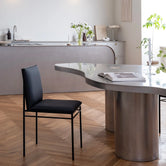 Contemporary dining space showcasing a By Crea Bella chair with a black seat and minimalist metal frame, positioned next to a uniquely shaped grey dining table with cylindrical bases. The background features a curved kitchen island and natural light. Ideal for modern interior design, sleek dining furniture, and stylish living spaces.