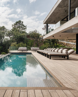 Modern outdoor poolside area featuring a row of Bazo Sunbed Loungers with cushioned headrests, set on a wooden deck beside a minimalist concrete villa surrounded by lush greenery.
