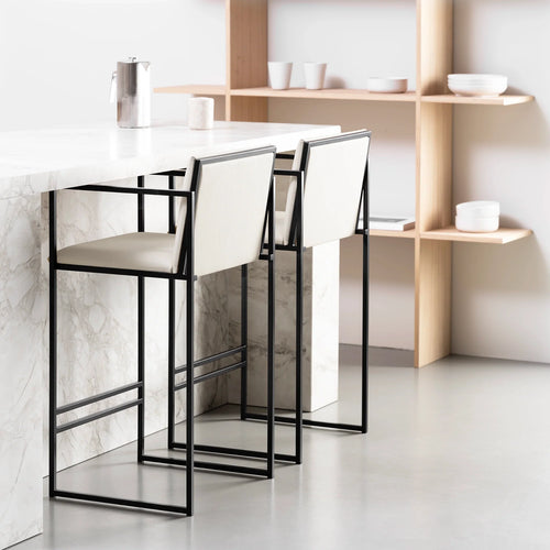 Two Bruno Bar Stools by by Crea with black steel frames and ivory vegan leather upholstery, placed at a white marble kitchen island in a Scandinavian-inspired interior with oak shelving.
