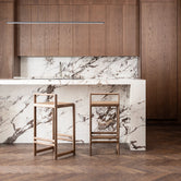 Stylish kitchen interior showcasing two By Crea wooden bar stools against a striking white marble island and backsplash, complemented by rich brown wooden cabinetry. Ideal for modern kitchen design inspiration, luxury home decor, and bespoke furniture.
