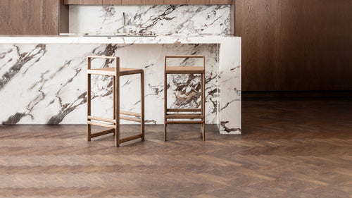 Pair of handcrafted Ebba Bar Stools in solid oak, styled in front of a dramatic marble waterfall kitchen island with dark wood cabinetry and herringbone flooring, showcasing Scandinavian minimalism and craftsmanship.