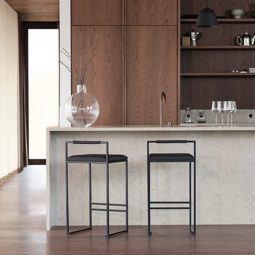 Minimalist kitchen island setup featuring two Freja Bar Stools with black steel frames, black vegan leather seats, and Jade black oak armrests, set against a light stone counter and warm wood cabinetry.