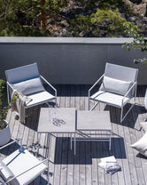Aerial view of an outdoor terrace featuring two by Crea Kent lounge chairs with white metal frames and plush light grey cushions, alongside a modular by Crea Anton lounge table with two light grey square tops, set on a weathered wood plank deck surrounded by greenery.