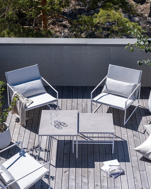 Aerial view of an outdoor terrace featuring two by Crea Kent lounge chairs with white metal frames and plush light grey cushions, alongside a modular by Crea Anton lounge table with two light grey square tops, set on a weathered wood plank deck surrounded by greenery.