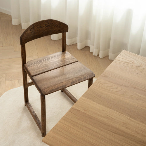 Karin dining chair in dark oak, showcasing elegant grain and soft edges, placed beside a wooden dining table.