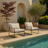 Two Kenny outdoor lounge chairs by Crea with black frames and cream cushions, styled poolside in a Mediterranean garden setting.