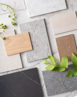 Flatlay of Scandinavian material swatches featuring oak wood in light and medium tones, and Dekton stone surfaces in warm neutrals, soft greys, and deep charcoal, styled with green leaves on a marble backdrop.
