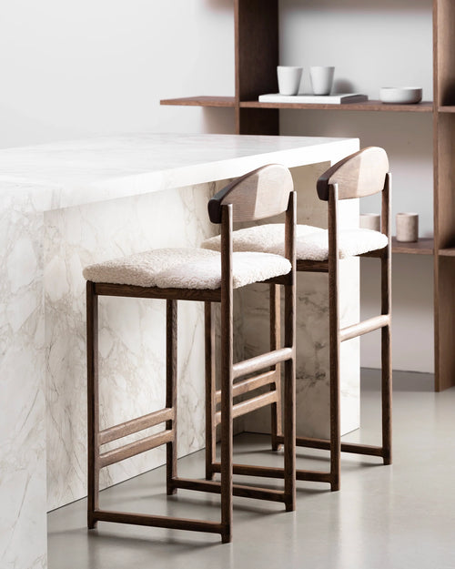 Contemporary kitchen featuring two Ted bar stools made of dark oak with curved wooden backs and soft white boucle cushions, pulled up to a veined white marble kitchen island, with wooden shelving in the background.