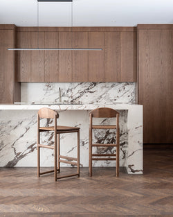Contemporary kitchen with custom dark wood cabinetry and herringbone parquet flooring, showcasing two Ted bar stools in dark oak wood with natural grain and curved backrests, pulled up to a luxurious white marble kitchen island with dark veining.