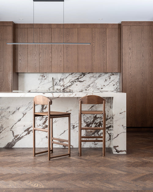Contemporary kitchen with custom dark wood cabinetry and herringbone parquet flooring, showcasing two Ted bar stools in dark oak wood with natural grain and curved backrests, pulled up to a luxurious white marble kitchen island with dark veining.