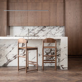 Two By Crea Ted bar stools crafted from solid dark oak, set in a luxurious kitchen with a white marble island and backsplash, and rich brown wooden cabinetry. Perfect for showcasing high-end Scandinavian design, sustainable furniture, and elegant kitchen interiors.