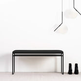 Front view of the Wilma bench in black steel frame with black cushion, styled with Flos IC S2 pendant lights in black, set against a minimalistic white interior.