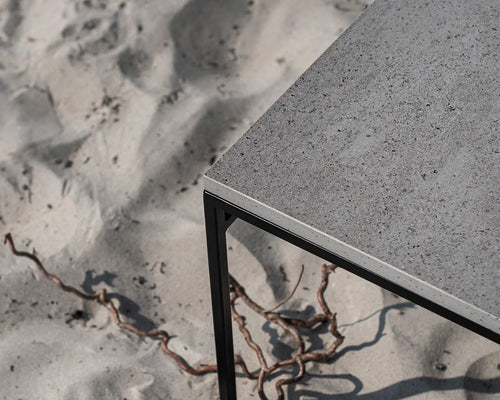 Detail view of the Wilma outdoor bench showing its black powder-coated steel frame and grey Dekton stone top, set against a sandy beach backdrop.