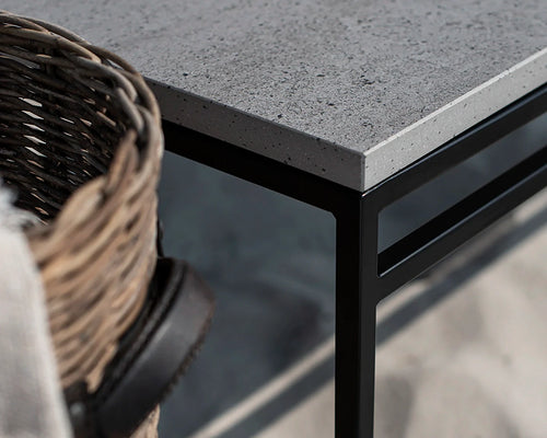 Detail of the Wilma outdoor bench featuring a black steel frame and grey Dekton stone top, styled with a wicker basket on a sandy beach setting.