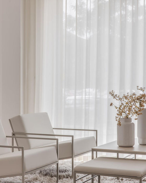 Minimalist lounge setup featuring two ivory-colored steel-framed armchairs and a matching ottoman from by Crea, styled with a low table and ceramic vases in a soft, monochrome room with sheer white curtains.