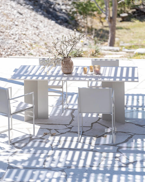 Scandinavian-inspired outdoor setup featuring the Ann dining table in beige by Crea, surrounded by white-framed chairs on a natural stone patio, styled with a ceramic vase and spring blossoms.