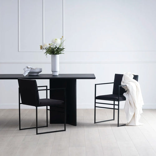 Ann dining table in black oak with two modern chairs and a vase of flowers in a minimalist setting.