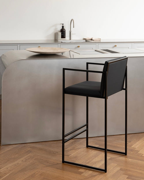 Close-up of a Bruno bar stool with black vegan leather seat and back, bold black metal frame with square armrests, next to a curved grey kitchen island with a gold faucet and wooden bowl on parquet flooring.