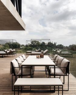 Minimalist outdoor dining area with the Diana Outdoor Table in Caesarstone, surrounded by black-framed armchairs with soft beige cushions, and matching bench seating on a wooden deck with panoramic views.