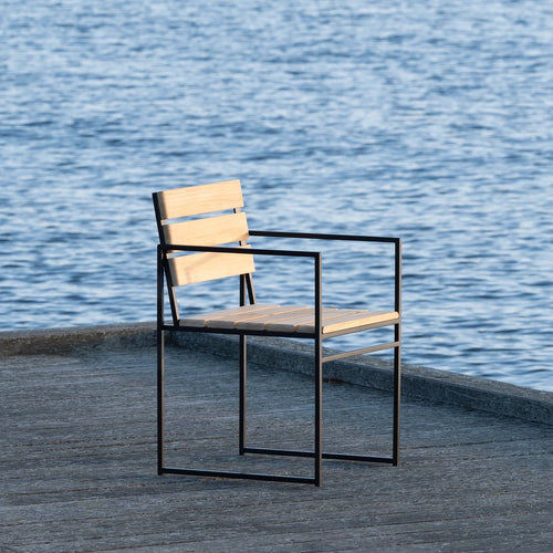 Elsy Outdoor Chair with armrests by by Crea in teak and black steel frame, pictured dockside against still blue water. Handmade Scandinavian outdoor furniture for modern and sustainable coastal living.