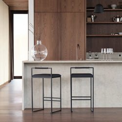 Minimalist kitchen island setup featuring two Freja Bar Stools with black steel frames, black vegan leather seats, and Jade black oak armrests, set against a light stone counter and warm wood cabinetry.