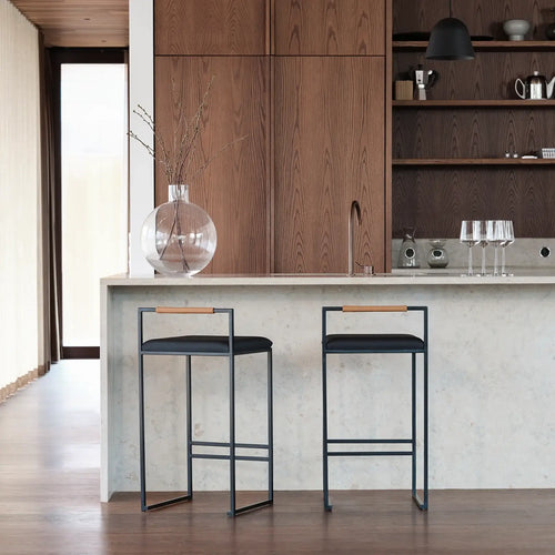 Freja bar stools in black leather at a modern kitchen island with a wooden backdrop and decorative vase.