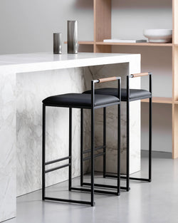 Freja bar stools by by Crea featuring black vegan leather cushions and Coffee Bean wooden backrests, styled at a modern marble kitchen island.