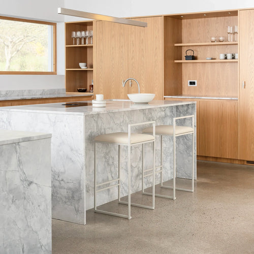 Freja Bar Stools in beige with minimalist frames, positioned at a grey-veined marble kitchen island in a light oak kitchen with open shelving.