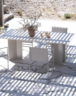 Sunny outdoor dining area featuring a sleek by Crea Ann rectangular outdoor table and matching chairs in a light beige or off-white finish, with sturdy block legs, placed on an irregularly paved stone patio, with strong striped shadows from an overhead pergola or trellis, and a vase of white blossoms as a centerpiece.