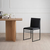 Front view of the Isabell Dining Chair from by Crea, featuring a black vegan leather seat and backrest with a minimalist black steel frame, styled next to a solid oak Ann dining table.