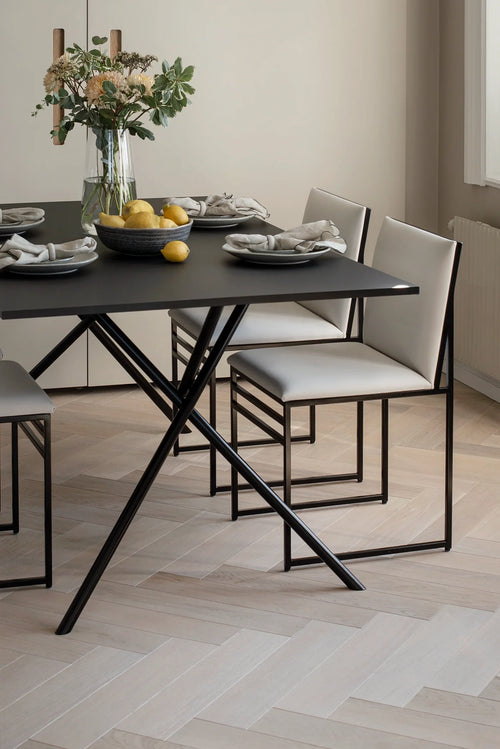 Melker Dining Table by by Crea with black X-frame base and matte top, paired with Isabell Dining Chairs in cream vegan leather and black steel frames in a minimalist Scandinavian dining room.