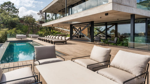 Modern poolside terrace featuring Kenny Outdoor Lounge Chairs with soft beige cushions and a row of Bazo Sunbed Loungers in black frames, arranged on a spacious wooden deck surrounding a luxury swimming pool.