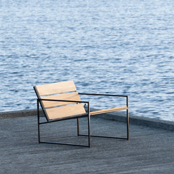 Kenny Lounge Chair in teak wood and black steel frame by by Crea, placed on a wooden dock beside still blue water. Showcasing sleek Scandinavian outdoor design, perfect for modern lakeside or coastal settings.