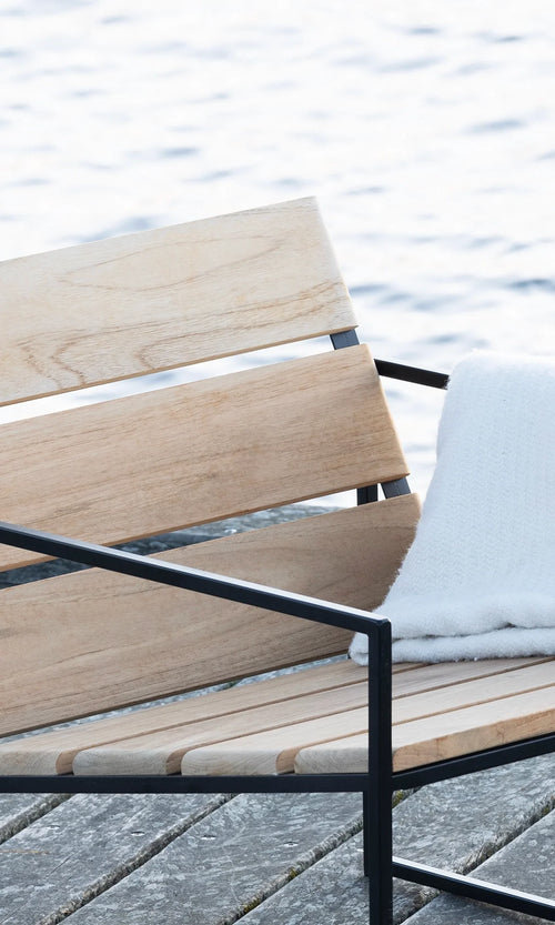 Kenny Outdoor Lounge Chair in teak wood and black frame by by Crea, shown dockside by the water with a soft white throw. Scandinavian design perfect for serene coastal settings, handmade in Sweden.