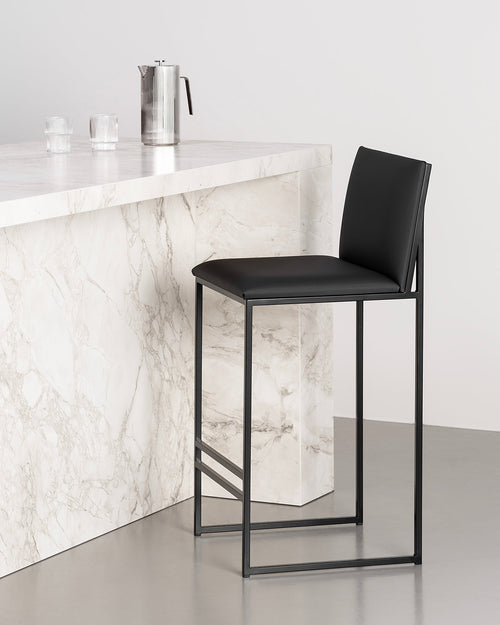 by Crea Philip bar stool in an all-black design with black vegan leather seat and back, and sleek black metal base, shown against a white marble kitchen island with a coffee pot and glasses.