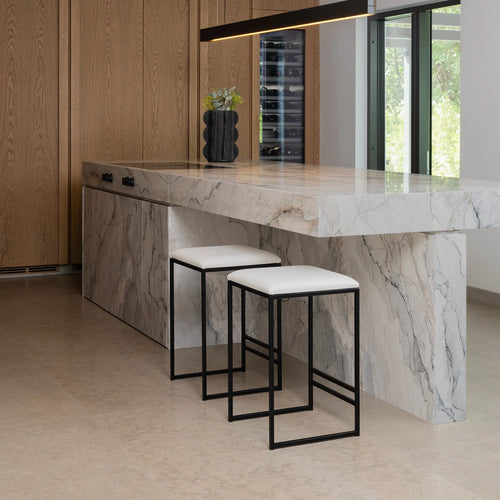 Svea Bar Stools with black powder-coated frames and ivory vegan leather seats, placed at a sculptural marble waterfall kitchen island in a modern interior with oak cabinetry and wine storage.