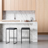 Two By Crea Svea all-black bar stools with sleek metal frames and dark seats, positioned at a white marble kitchen island, with light wooden cabinetry and a marble backsplash in the background. Ideal for modern minimalist kitchen designs and contemporary furniture.
