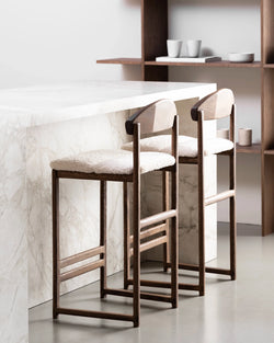 Contemporary kitchen featuring two Ted bar stools made of dark oak with curved wooden backs and soft white boucle cushions, pulled up to a veined white marble kitchen island, with wooden shelving in the background.