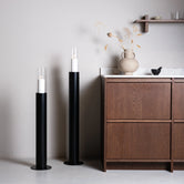 Scandinavian black floor candleholders with glass cylinders beside a wooden cabinet in a stylish room setting.