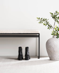 Minimalist entryway with the Wilma bench by Crea in a black steel frame and ivory bouclé cushion, black ankle boots tucked beneath, and a large stone vase with greenery beside it.