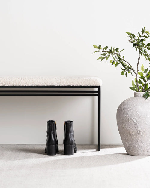 Minimalist entryway with the Wilma bench by Crea in a black steel frame and ivory bouclé cushion, black ankle boots tucked beneath, and a large stone vase with greenery beside it.