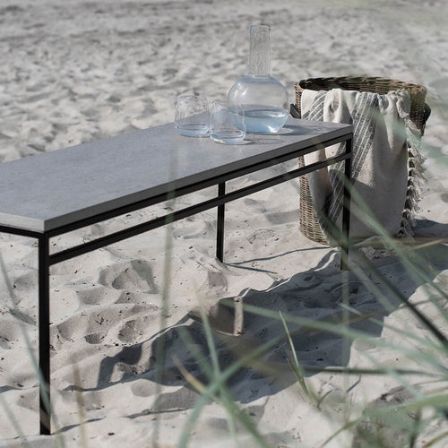 Wilma outdoor bench with sleek Keon dekton top on the beach, complemented by glassware and a woven basket.