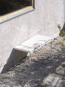 Wilma outdoor bench by by Crea in white frame with a light Dekton stone top, placed on gravel against a stucco wall in a minimal garden setting.