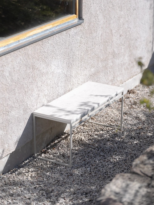 Wilma outdoor bench by by Crea in white frame with a light Dekton stone top, placed on gravel against a stucco wall in a minimal garden setting.